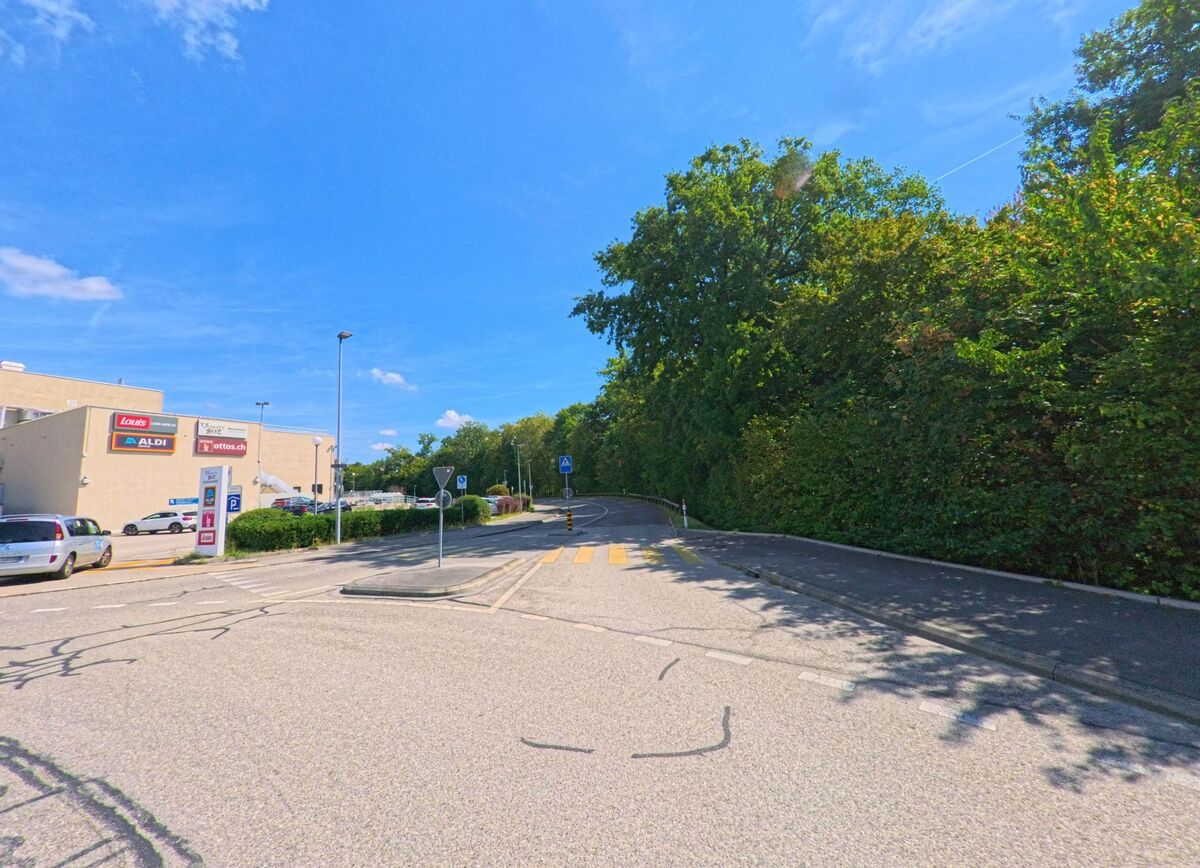 Am Rand von Etoy (1163, Kanton Waadt) liegt ein Einkaufszentrum mit ALDI, daneben führt eine Strasse mit Zebrastreifen in ein von Bäumen gesäumtes Gebiet.