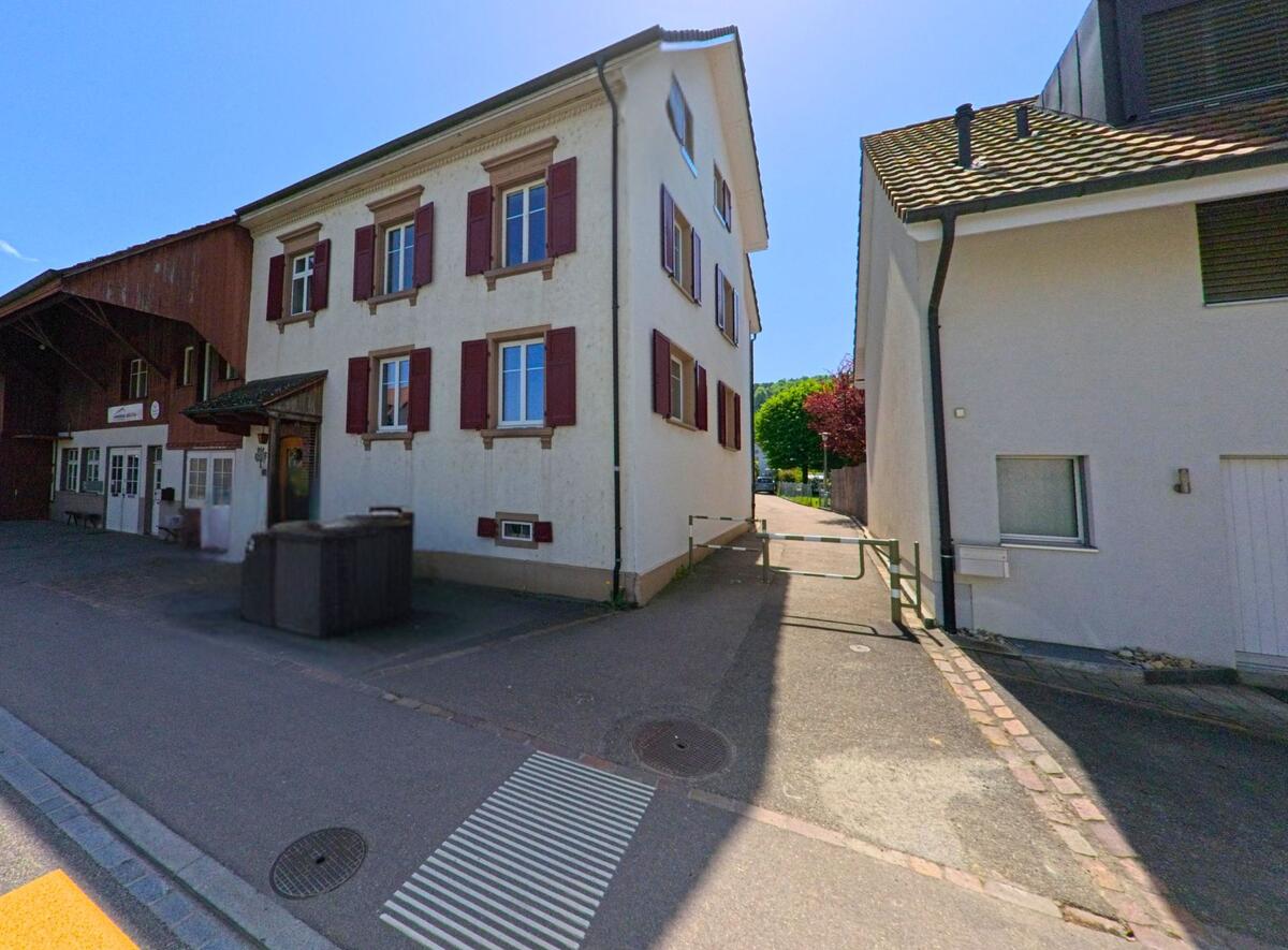 In Ettingen (4107, Kanton Baselland) zeigt sich ein Gebäude mit einem traditionellen Holzbau sowie modernen Elementen. Ein blauer PKW steht davor, während die Straße ruhig verläuft.