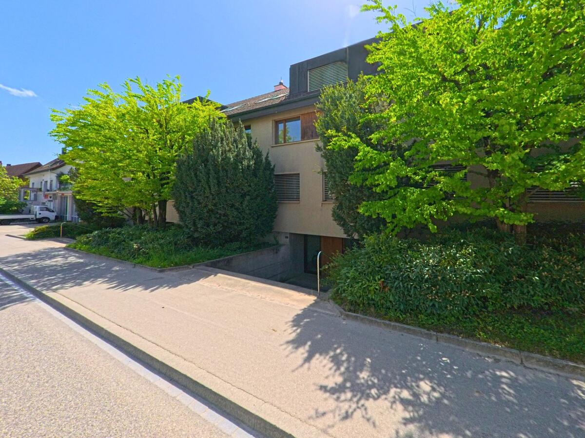 In Ettingen (4107, Kanton Baselland) steht ein modernes Mehrfamilienhaus, umgeben von grünen Bäumen und gut gepflegtem Grünstreifen entlang einer ruhigen Strasse.