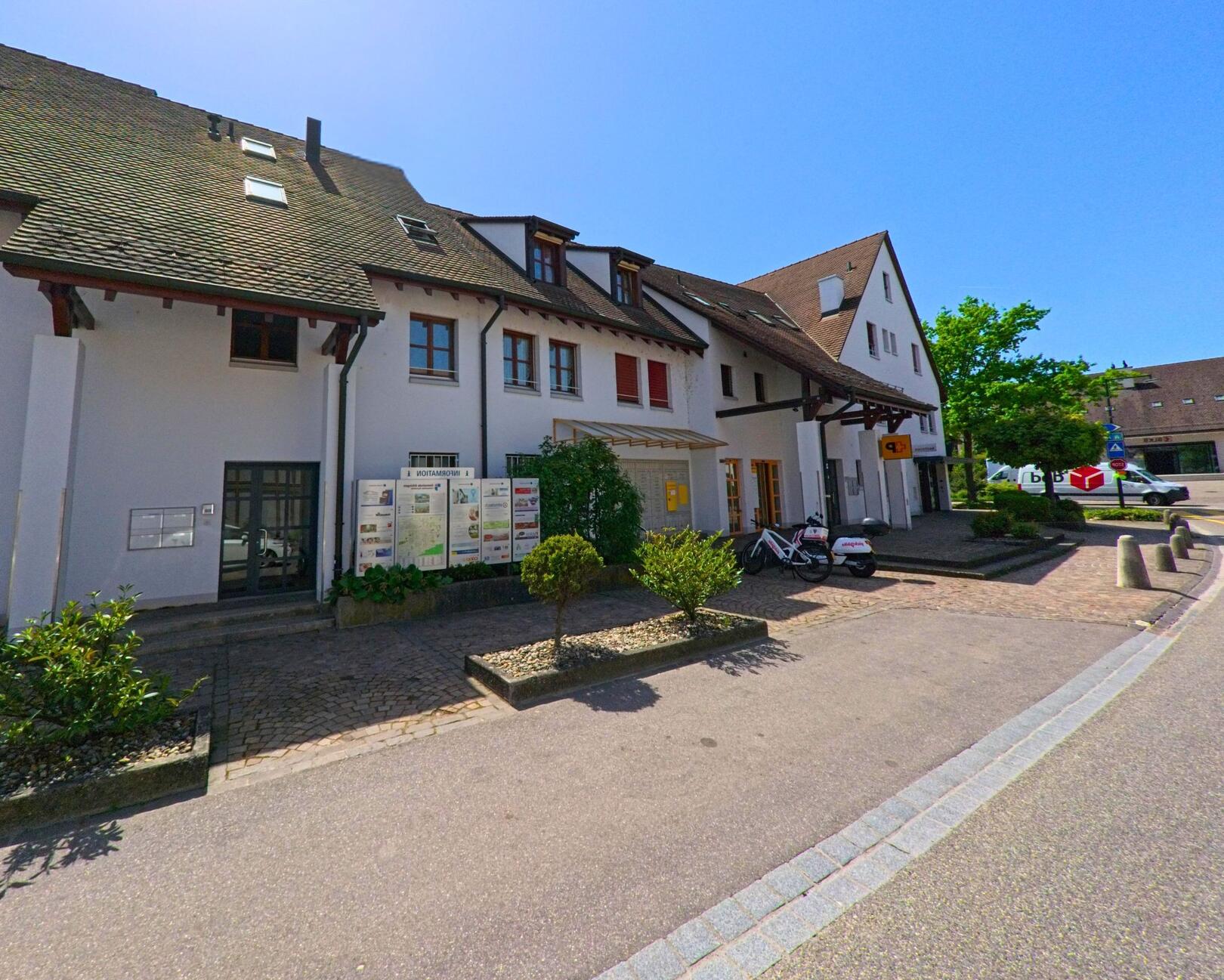 In Ettingen (4107, Kanton Baselland) befindet sich ein mehrgeschossiges Wohn- und Geschäftshaus mit modernen Fensterläden und einem ausgeschilderten Privatparkplatz.