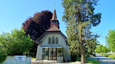 Eine kleine Kirche mit Turm und modernem Eingangsbereich steht zwischen grossen Baeumen in St. Gallen (9000, Kanton Widnau) an einer ruhigen Strasse.