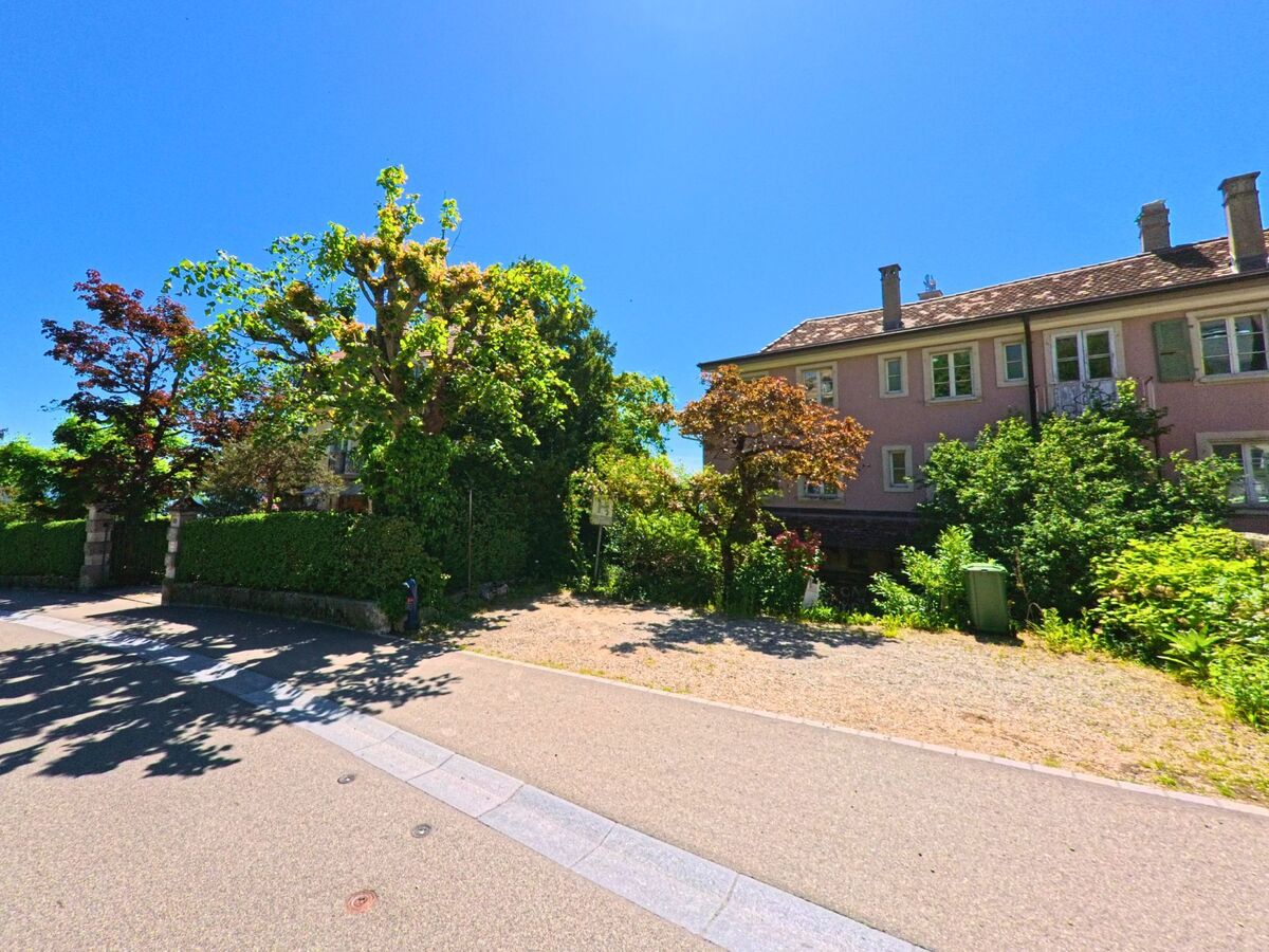 In Evilard (2533, Kanton Bern) stehen Wohnhäuser mit hellen Fassaden hinter üppig bewachsenen Vorgärten und schattenspendenden Bäumen.