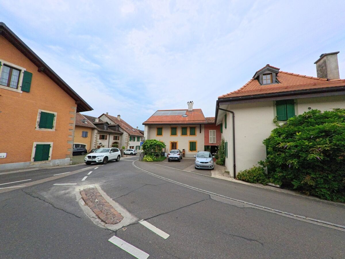Eine enge Strasse in Eysins (1262, Kanton Waadt), mit Häusern in verschiedenen Farben und parkenden Autos entlang der Kurve.