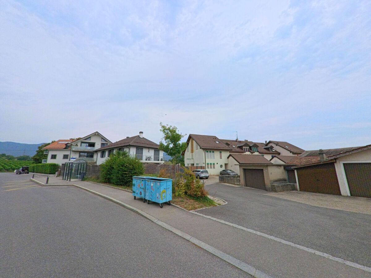 Eine ruhige Strasse in Eysins (1262, Kanton Waadt) zeigt Wohnhäuser und Garagen, mit einem bunten Müllcontainer auf dem Gehweg und Weinbergen im Hintergrund.