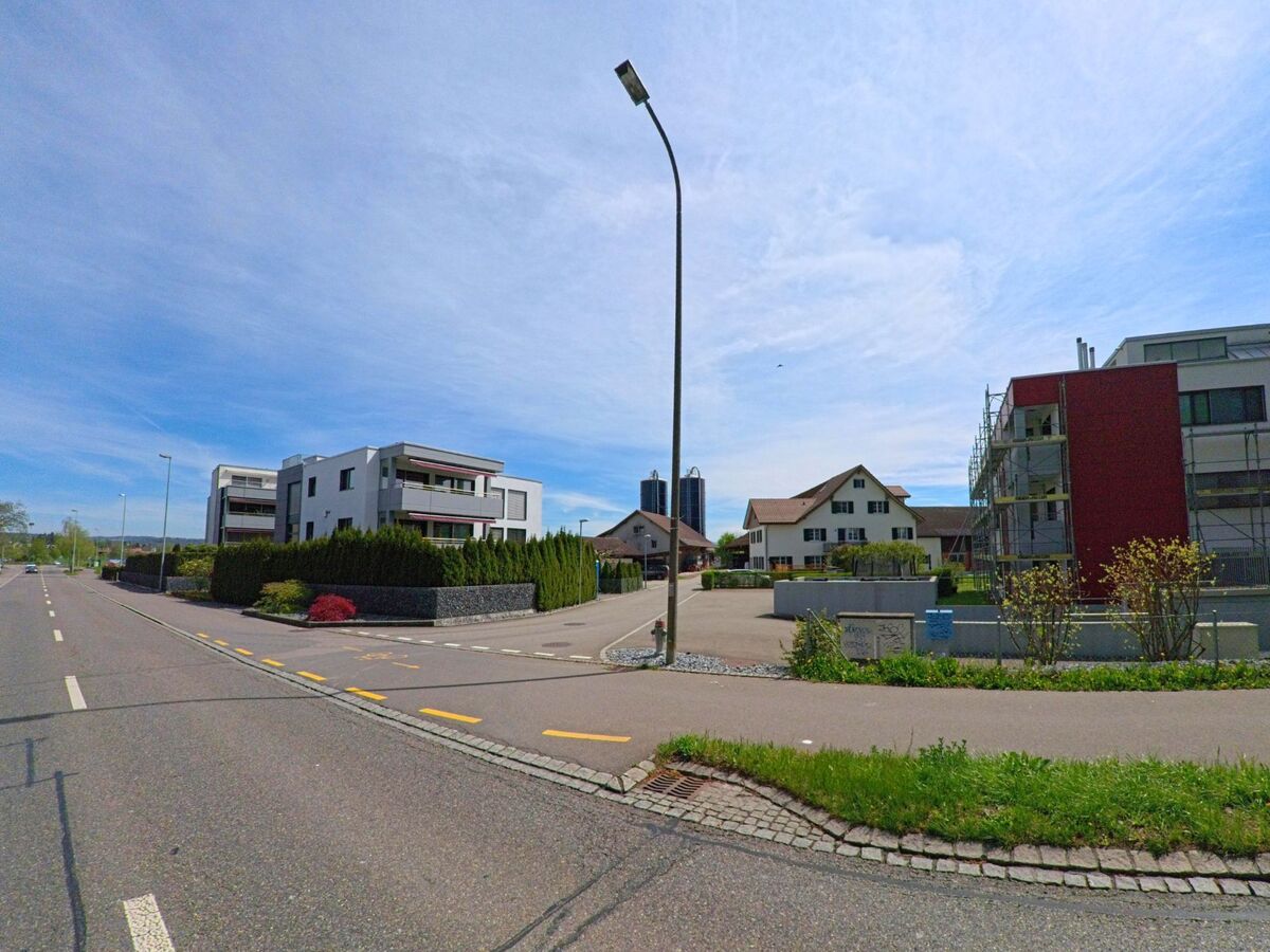 An einer breiten Strasse in Fällanden (8117, Kanton Zürich) stehen moderne Mehrfamilienhäuser und ein traditionelles Wohnhaus, flankiert von Gehwegen und Grünflächen.