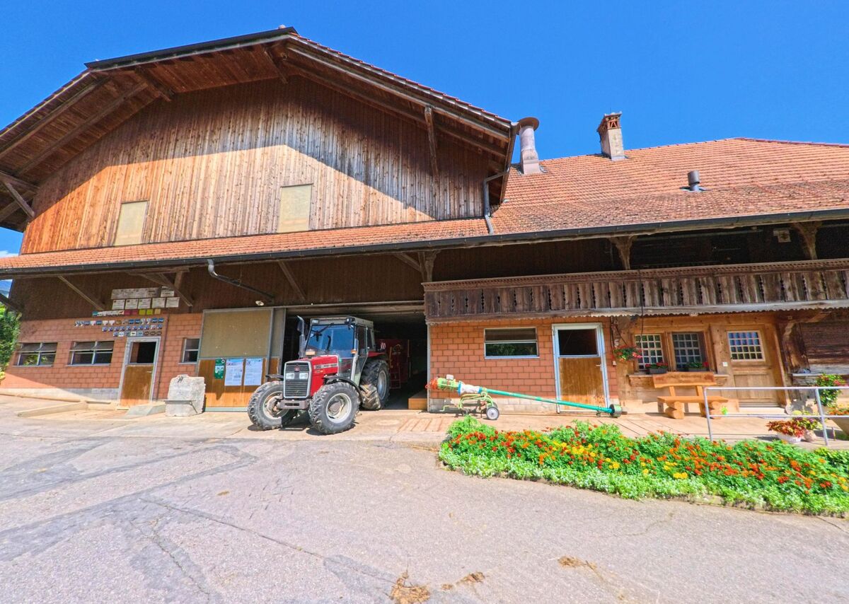 Ein grosser Bauernhof in Fahrni (3617, Kanton Bern) mit Backsteinfassade und Holzaufbau beherbergt einen roten Traktor in der offenen Halle, davor bluht ein farbenfrohes Blumenbeet.