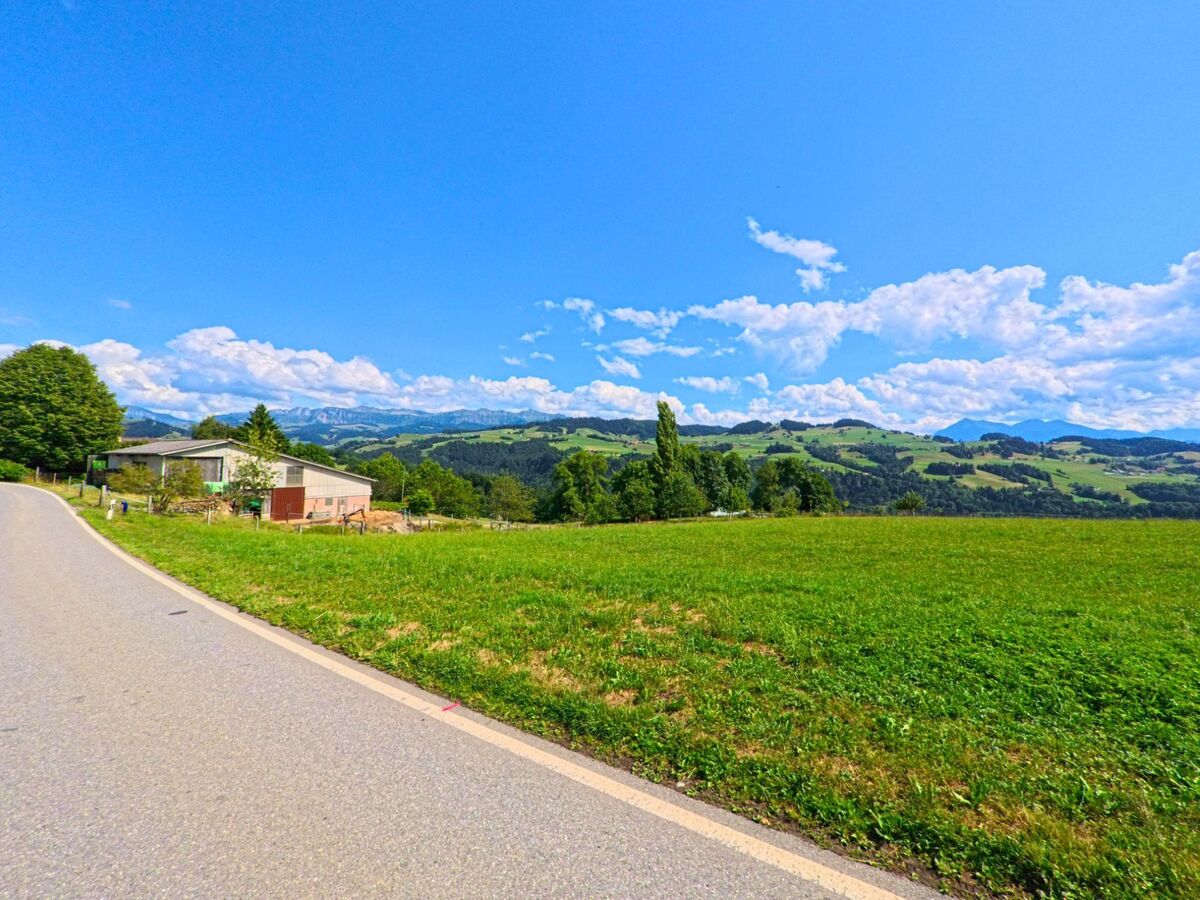 In Fahrni (3617, Kanton Bern) fuehrt eine schmale Landstrasse an einem einzelnen Nebengebaeude vorbei durch offene Felder mit Blick auf die bewaldeten Huegel und entfernte Bergketten.