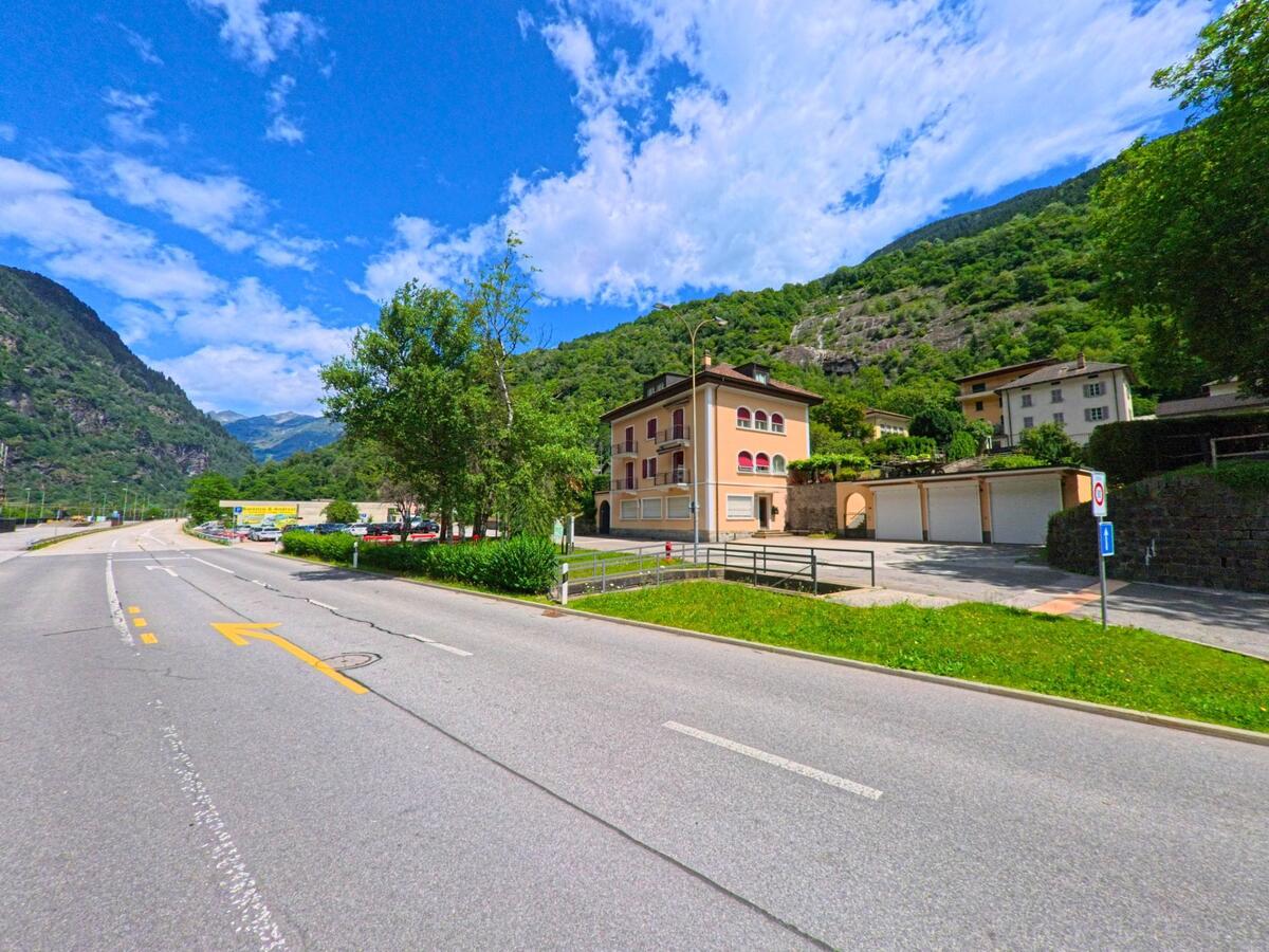 An einer breiten Strasse in Faido (6746, Kanton Tessin) stehen mehrgeschossige Wohnhäuser mit Balkonen vor einer bewaldeten Bergkulisse und blauem Himmel.