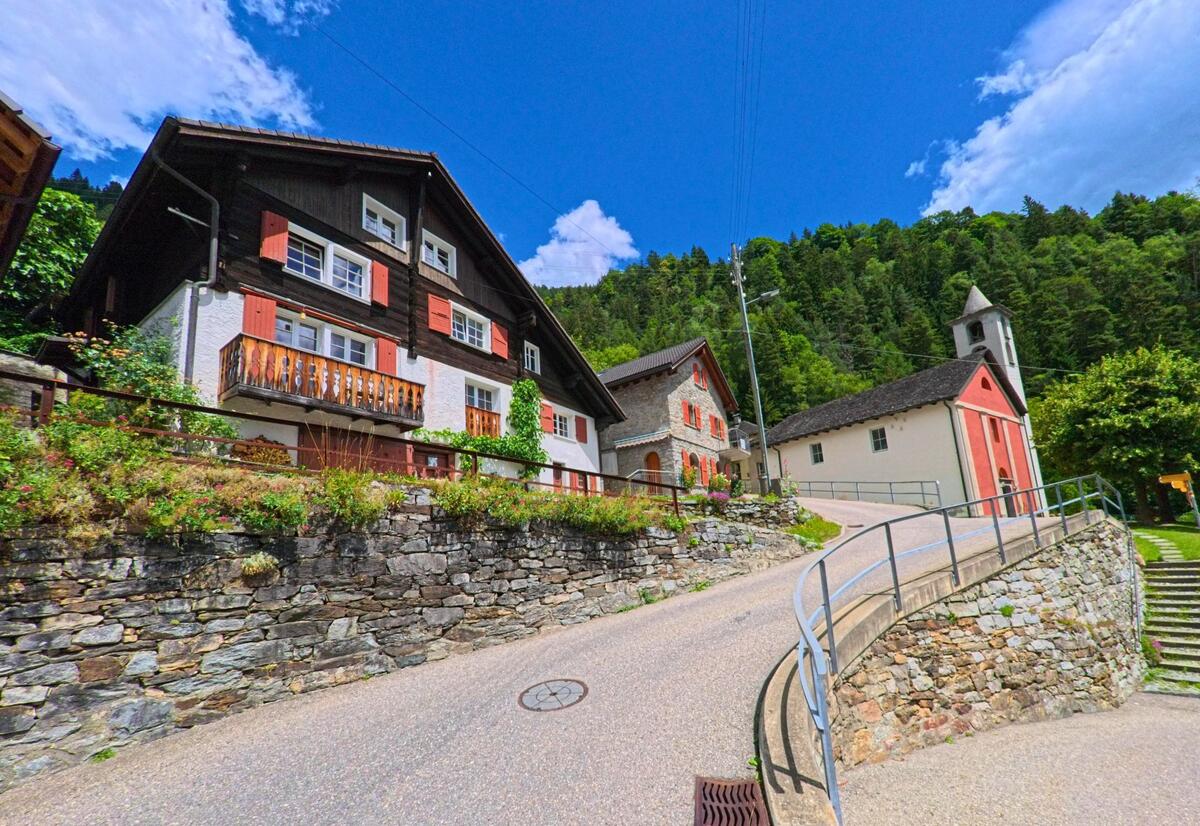 An einem Dorfhang in Faido (6748, Kanton Tessin) stehen ein Holzhaus mit roten Fensterläden und Steinhäuser an einer ansteigenden Strasse, dahinter eine kleine Kirche mit rotem Glockenturm vor bewaldeten Berghängen.