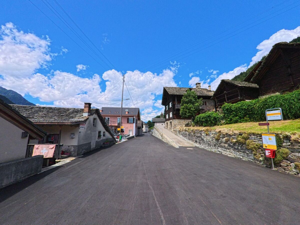 Eine schmale Dorfstrasse in Faido (6749, Kanton Tessin) fuehrt zwischen alten Steinhaeusern mit Schindeldaechern und gepflegten Gaerten den Hang hinauf.