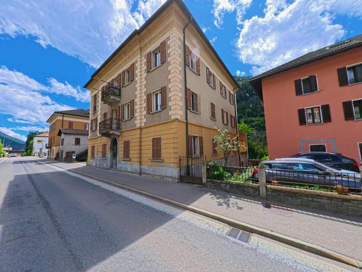 In einer sonnigen Strasse von Faido (6760, Kanton Tessin) stehen traditionelle Wohnhäuser mit farbigen Fassaden und geschlossenen Fensterläden vor einem klaren Himmel.