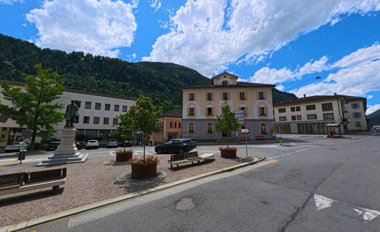 Auf dem zentralen Platz von Faido (6763, Kanton Tessin) steht eine Statue vor dem Gemeindehaus, umgeben von gepflasterten Wegen, Baenken und Blumenkuebeln mit Blick auf den bewaldeten Hang.