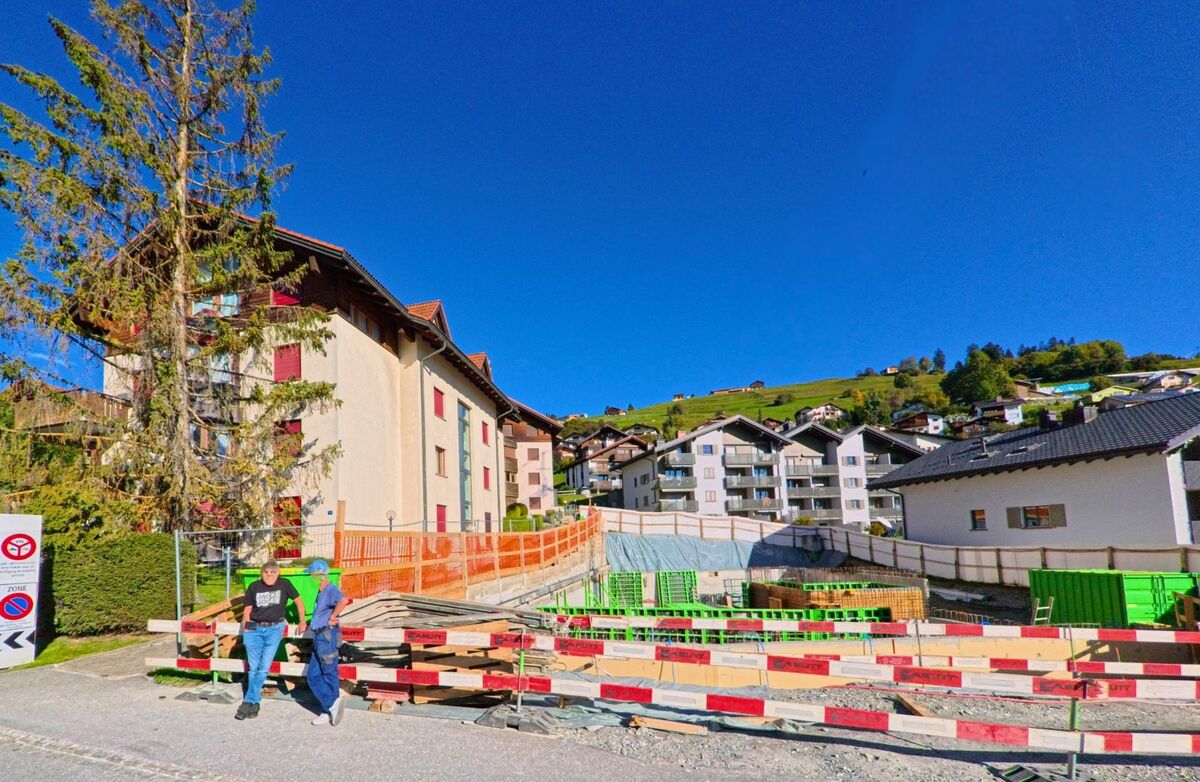 In Falera (7153, Kanton Graubuenden) befindet sich eine Baustelle zwischen modernen Wohnhaeusern am Hang, waehrend zwei Maenner am Rand des Baugebiets stehen.