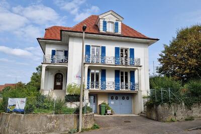 Ein dreigeschossiges Haus mit rotem Dach und blauen Fensterlaeden steht in Schoenenbuch (4124, Kanton Baselland) an einer ruhigen Strasse.