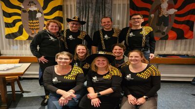 In Ibach (6438, Kanton Schwyz) praesentiert sich der Vorstand der Fasnachtsgesellschaft in einheitlicher Kleidung vor farbigen Fasnachtsmotiven in einem Innenraum.