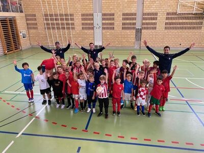 In einer Turnhalle in Risch (6343, Kanton Zug) versammeln sich zahlreiche Kinder des FC Rotkreuz mit ihren Trainern zu einem fröhlichen Gruppenfoto mit erhobenen Armen.