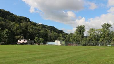 Ein gepflegter Fussballplatz mit Toren und Vereinsgebaeuden liegt am Rand von bewaldeten Huegeln in Schleitheim (8226, Kanton Schaffhausen).