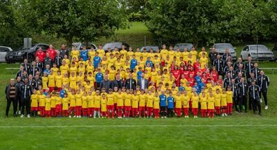 In Wollerau (8832, Kanton Schwyz) versammeln sich zahlreiche Kinder und Erwachsene des FC Wollerau in gelben und roten Trikots zu einem grossen Vereinsfoto auf dem Rasen.