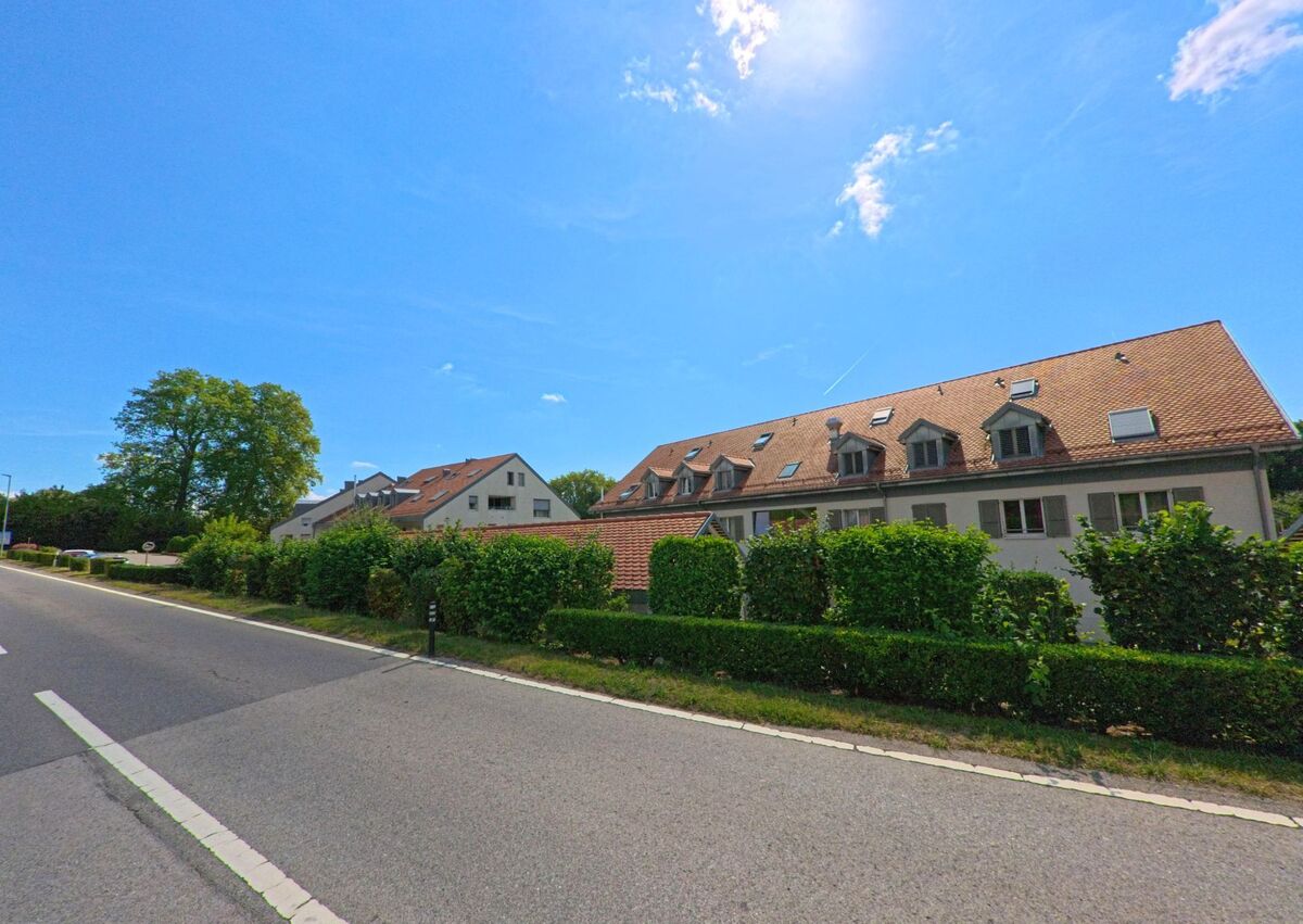 Mehrfamilienhäuser mit roten Dächern stehen in Féchy (1173, Kanton Waadt) entlang einer Strasse, gesäumt von gepflegten Hecken.