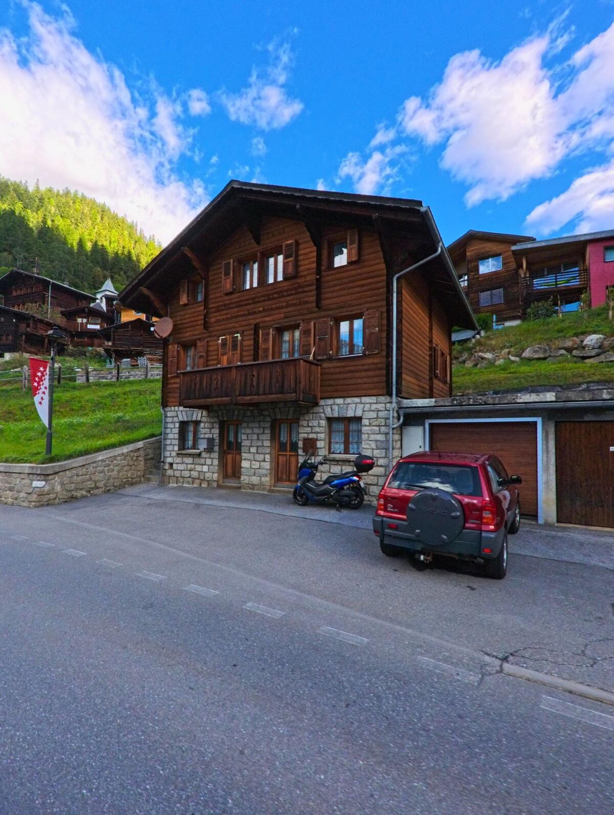 Ein traditionelles Chalet in Ferden (3916, Kanton Wallis) mit Steinsockel und Holzbalkon steht am Strassenrand, davor sind ein Auto und ein Motorroller geparkt.
