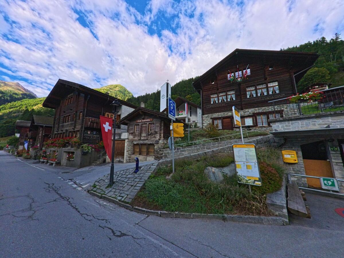 Im Dorfkern von Ferden (3916, Kanton Wallis) stehen traditionelle Holzhäuser, davor befinden sich eine Bushaltestelle und eine Schweizer Fahne.