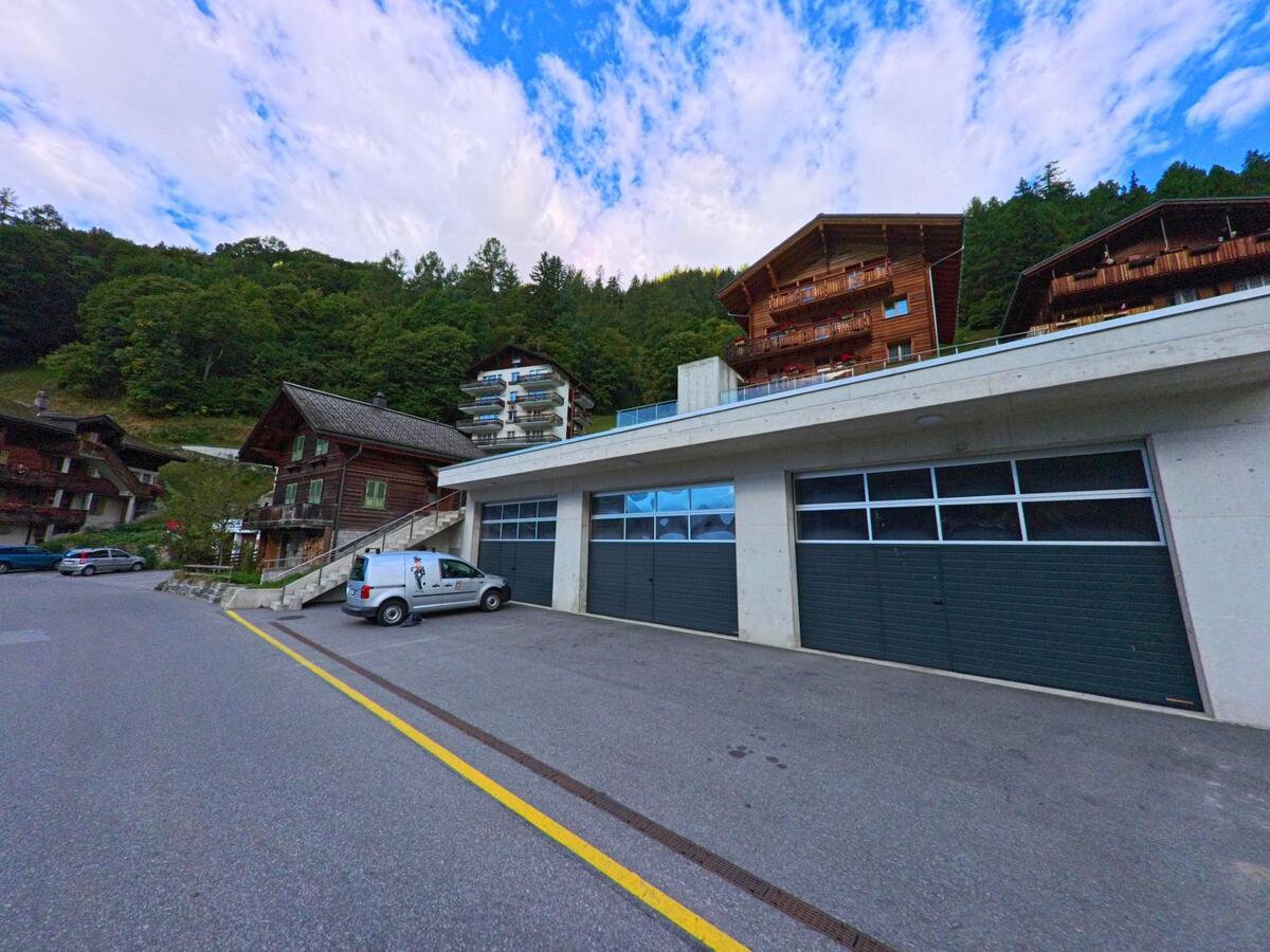 Am Ortsrand von Ferden (3916, Kanton Wallis) stehen moderne Garagen neben traditionellen Holzhäusern und Chalets mit Balkonen am bewaldeten Hang.