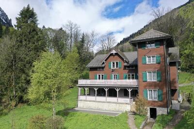 Das Ferienheim Morgenholz in Glarus Nord (8757, Kanton Glarus) ist ein mehrstöckiges Holzgebäude mit grünen Fensterläden, Balkon und Turm, umgeben von Wiesen, Bäumen und bewaldeten Berghängen.