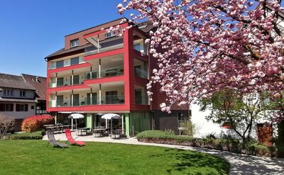 In Berlingen (8267, Kanton Thurgau) steht ein modernes rotes Hotelgebäude mit Balkonen, davor blüht ein Kirschbaum in voller Pracht.
