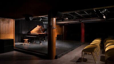 Im Konzertsaal der Ferme-Asile in Sion (1950, Kanton Wallis) steht ein Flügel auf einer beleuchteten Bühne vor schwarzen Vorhängen, umgeben von gelben Stühlen für das Publikum.