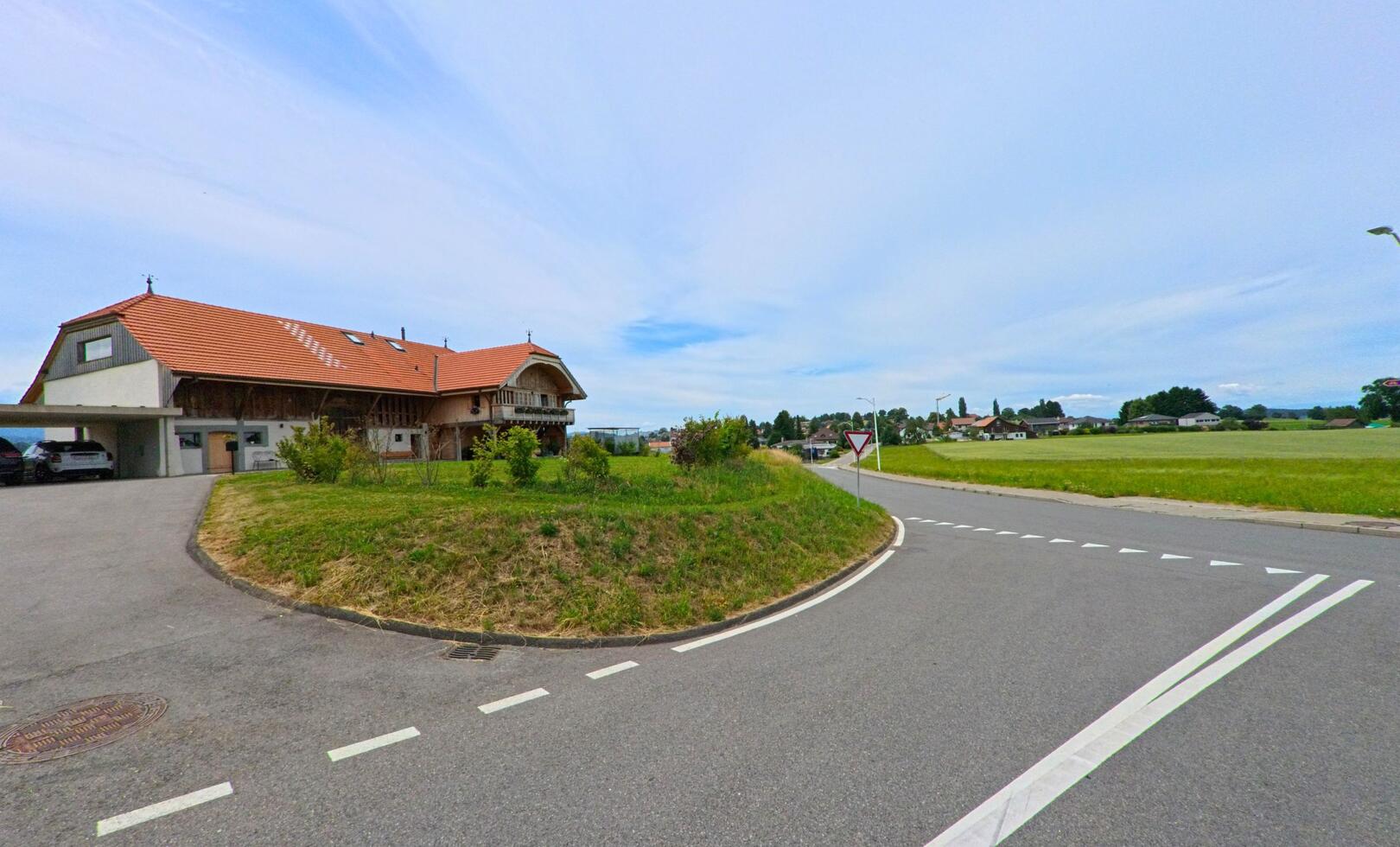 In Ferpicloz (1724, Kanton Freiburg) steht ein grosses Bauernhaus mit rotem Ziegeldach an einer Weggabelung, umgeben von Wiesen und wenigen Nachbarhäusern.