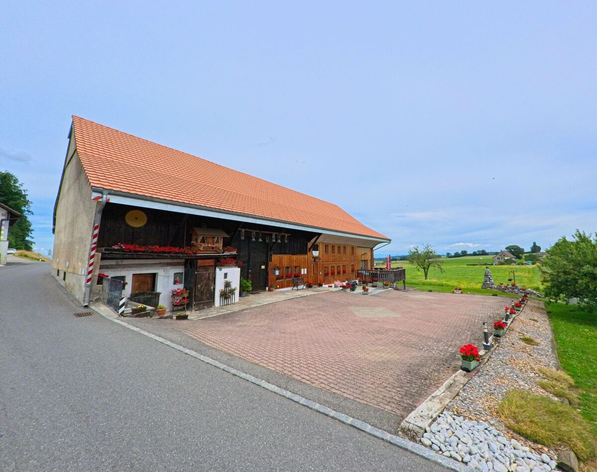 In Ferpicloz (1724, Kanton Freiburg) steht ein geschmückter Bauernhof mit rotem Ziegeldach und grossem Vorplatz, umgeben von Feldern und Obstbäumen.