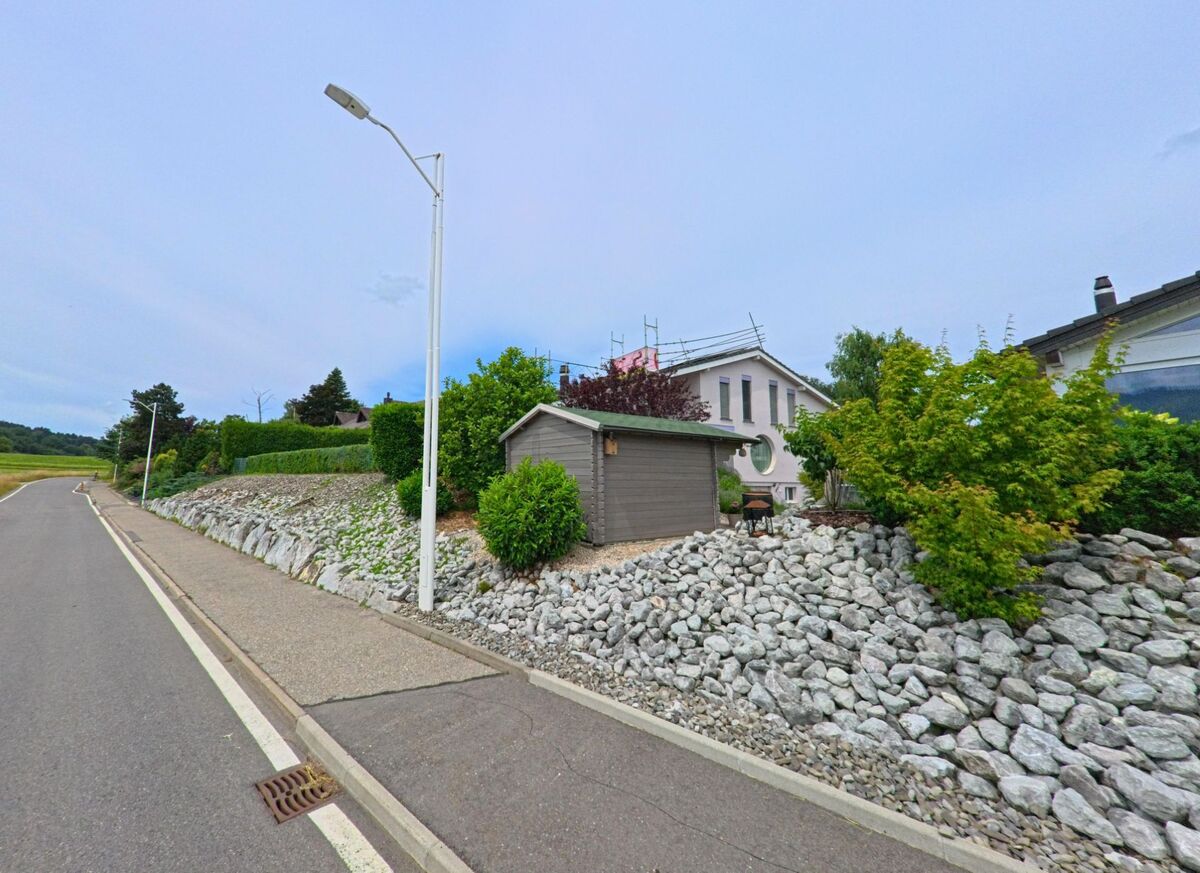 In Ferpicloz (1724, Kanton Freiburg) steht ein traditionelles Holzhaus mit Blumenbrunnen an einer Kurve, beschattet von einem grossen Laubbaum..