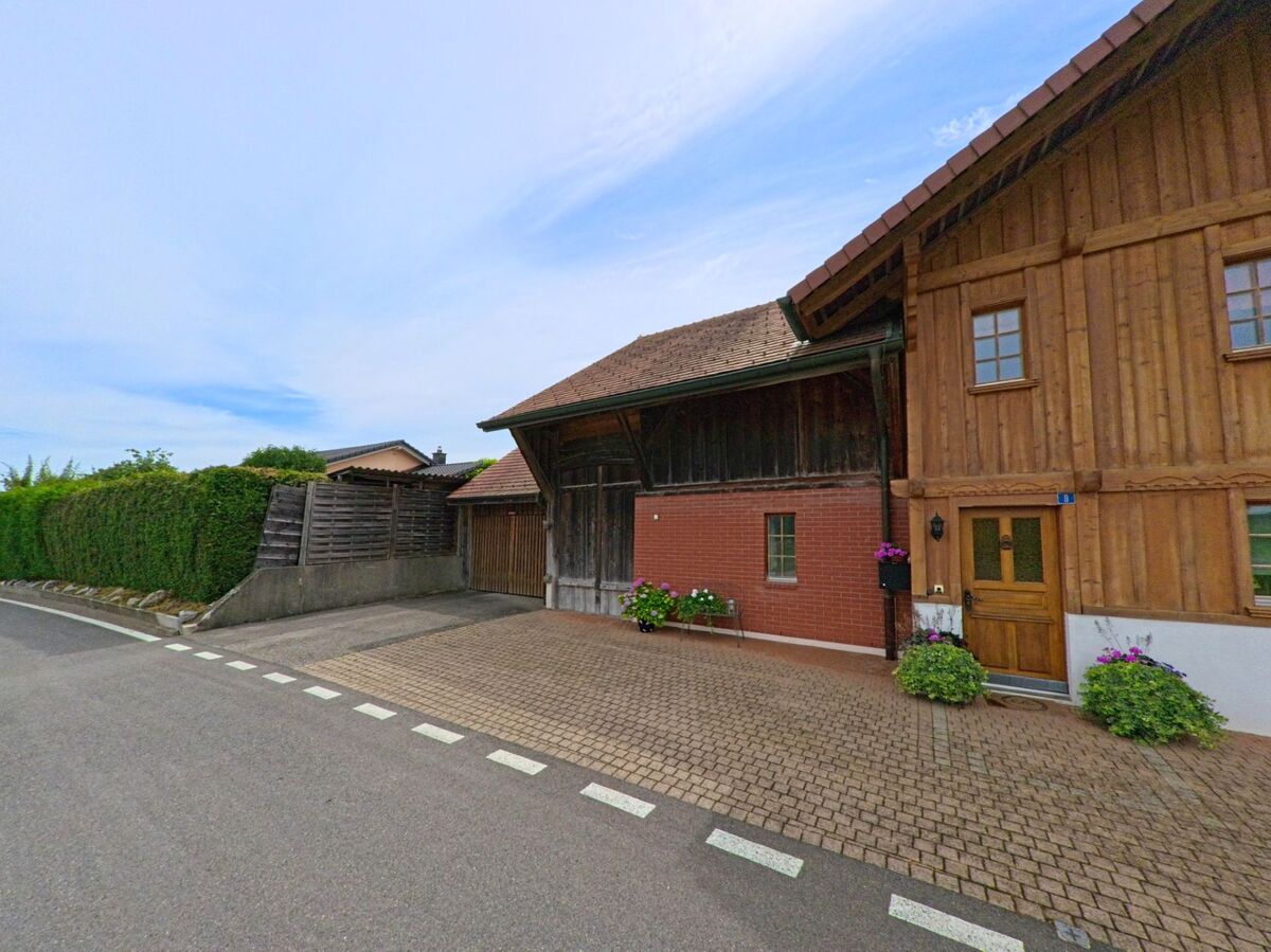 In Ferpicloz (1724, Kanton Freiburg) steht ein liebevoll gepflegtes Holzhaus mit Blumen am Eingang direkt an einer schmalen Strasse mit Hecken und Einfriedungen.