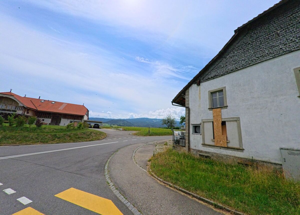 In Ferpicloz (1724, Kanton Freiburg) liegt ein leerstehendes Gebäude an einer Strassenkurve, gegenüber von einem renovierten Bauernhaus mit Blick auf die weite Landschaft.