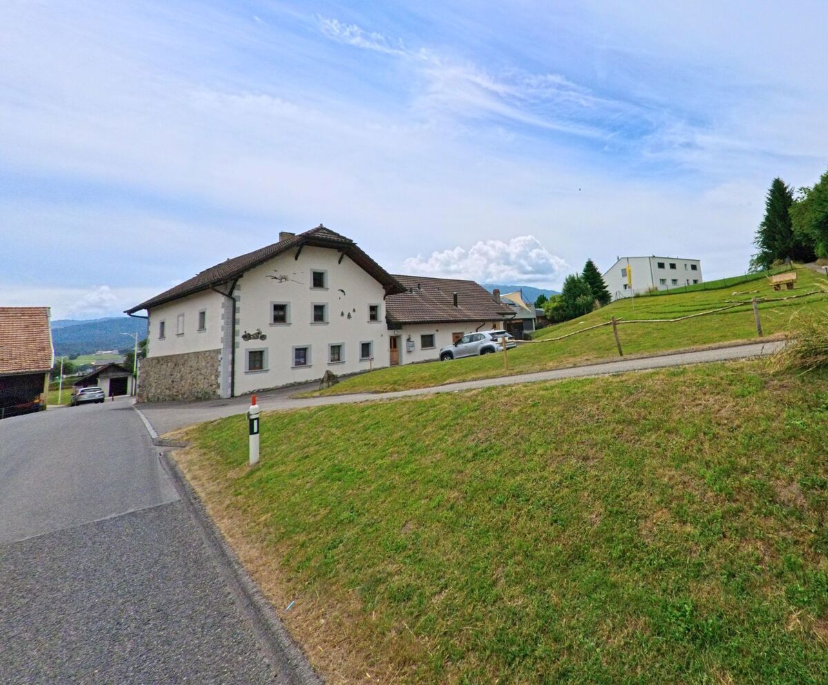 In Ferpicloz (1724, Kanton Freiburg) steht ein weiss verputztes Wohnhaus an einer ländlichen Strasse mit Blick auf eine Anhöhe mit Wald und modernen Gebäuden.