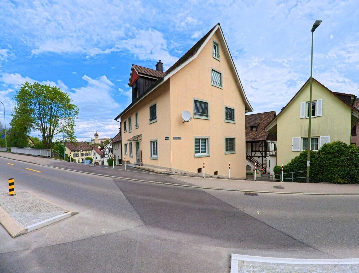  In Feuerthalen (8245, Kanton Zürich) führt eine Straße an einem gelben Gebäude vorbei, mit einem weiteren Haus im Hintergrund und der Kirche, die in der Ferne sichtbar ist.