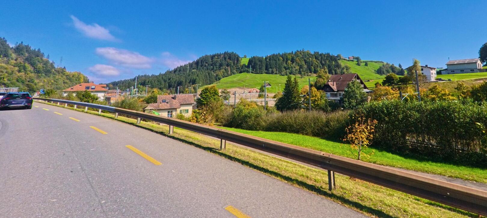 Auf einer kurvigen Landstrasse in [ORT] (PLZ [PLZ], [KANTON]) fahrt ein Auto entlang saftig gruener Wiesenlandschaft, waehrend sich im Hintergrund das bergige Terrain erhebt.