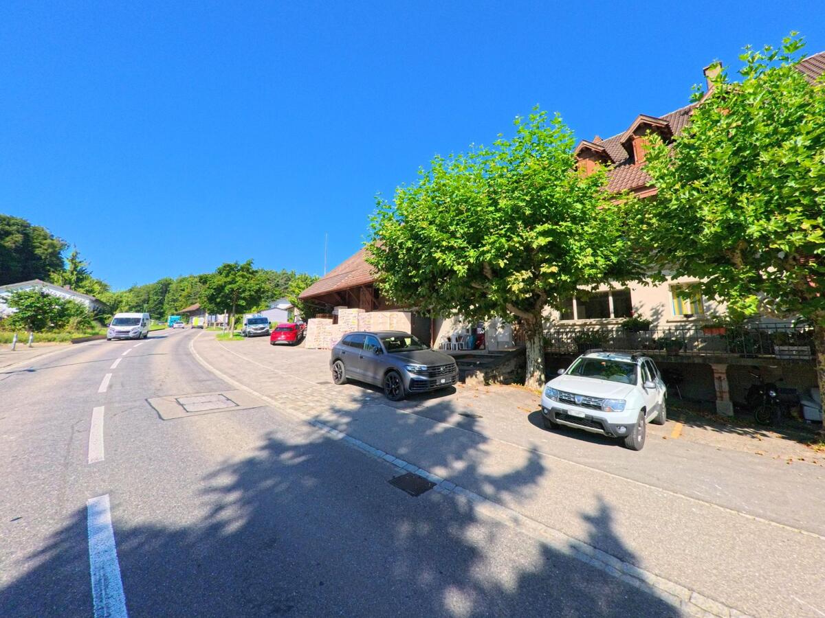 An der Hauptstrasse in Finsterhennen (2577, Kanton Bern) stehen grosse Gebäude und parkende Autos im Schatten von Baumreihen.
