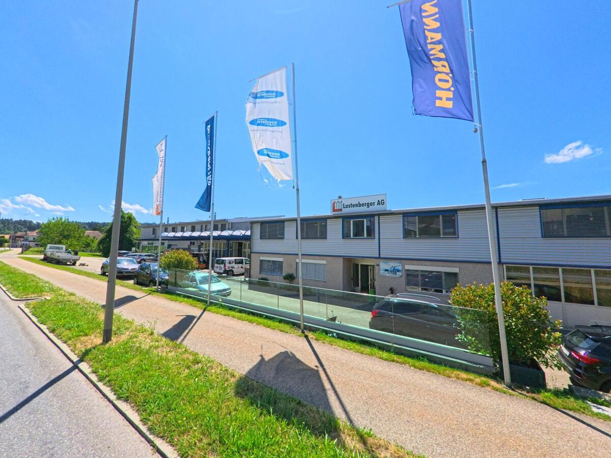 Das Gewerbegebaeude der Lustenberger AG steht in Fischbach (6145, Kanton Luzern) an einer Nebenstrasse, gesaeumt von wehenden Firmenflaggen und geparkten Fahrzeugen.