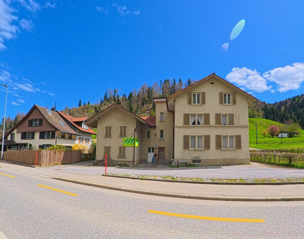 In Fischenthal im PLZ-Bereich 8497 im Kanton Zürich steht ein einfaches Wohnhaus mit Fensterläden an einer Hauptstrasse, umgeben von Hügeln und vereinzelten Bäumen.