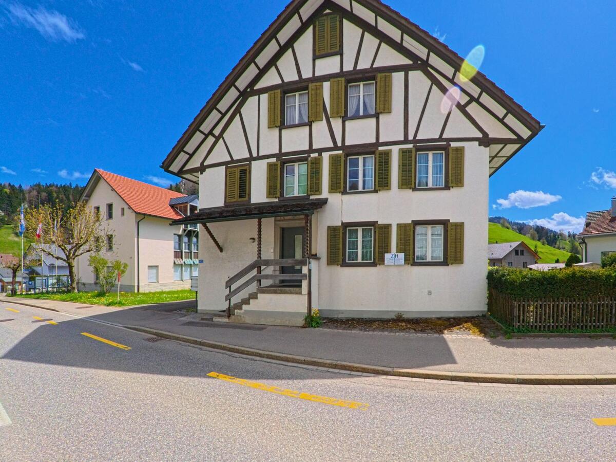 In Fischenthal im Kanton Zürich, PLZ 8497, steht ein traditionelles Fachwerkhaus mit grünen Fensterläden an einer leicht gebogenen Strasse vor hügeliger Landschaft.