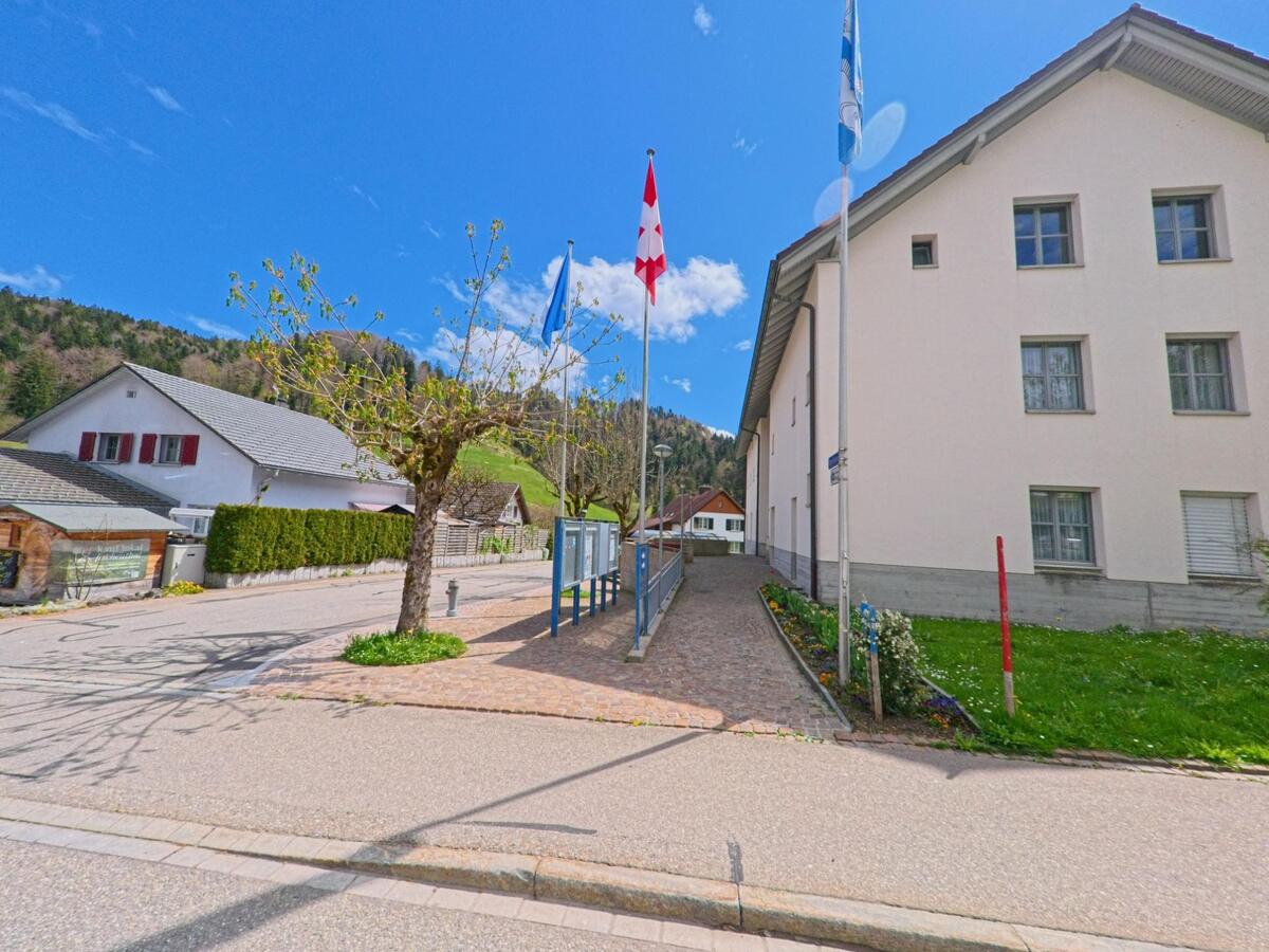 In Fischenthal im Kanton Zürich, PLZ 8497, steht ein öffentliches Gebäude mit Schweizer und Zürcher Fahnen sowie einem Infoboard an einem gepflasterten Weg im Ortszentrum.