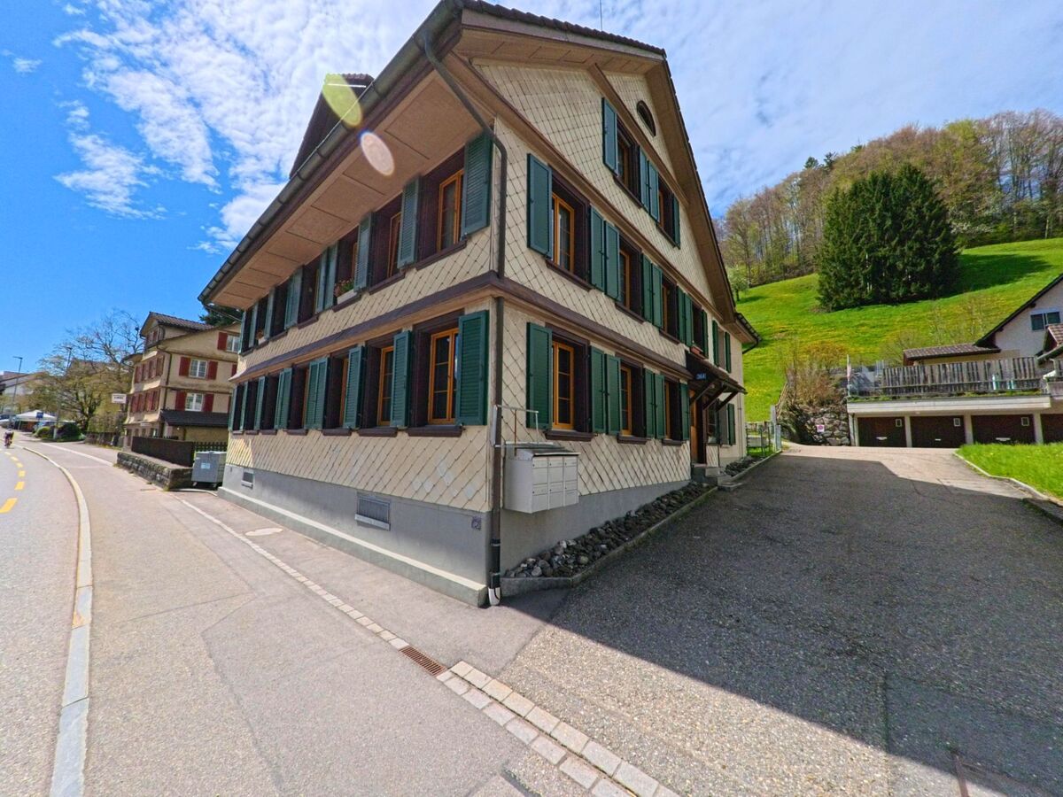  In Fischenthal im Kanton Zürich, PLZ 8497, steht ein traditionelles Wohnhaus mit grünen Fensterläden an einer leicht geschwungenen Strasse vor einem bewaldeten Hang.