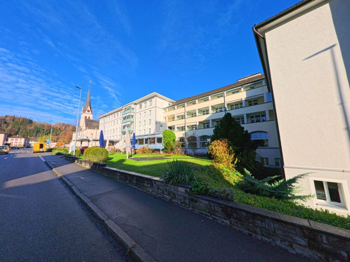 Entlang einer Strasse in Fischingen (8374, Kanton Thurgau) liegt eine gepflegte Grünanlage vor einem grossen Gebäudekomplex, im Hintergrund ragt ein Kirchturm auf.