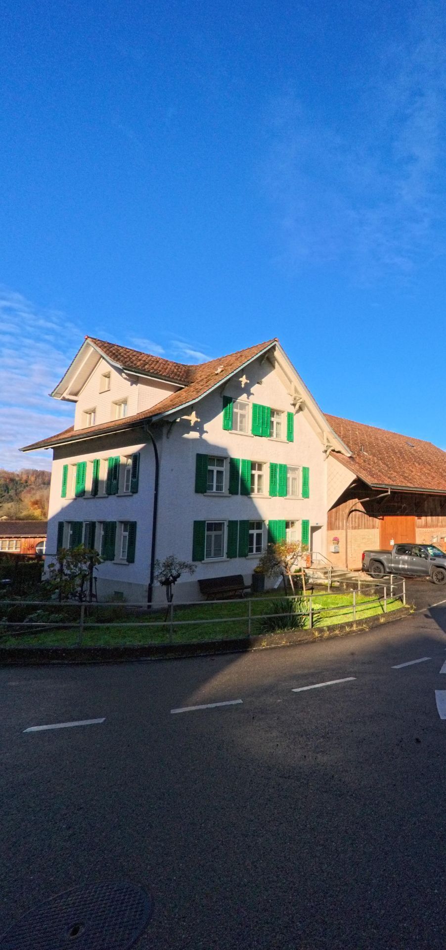 Ein weisses Wohnhaus mit grünen Fensterläden steht in Fischingen (8374, Kanton Thurgau) an einer Strasse neben einem Scheunengebäude.