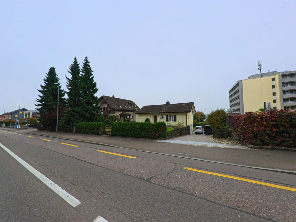 Entlang der Hauptstrasse in Fislisbach (5442, Kanton Aargau) stehen Einfamilienhäuser mit Vorgärten und hohen Tannen, im Hintergrund sind Mehrfamilienhäuser zu sehen.