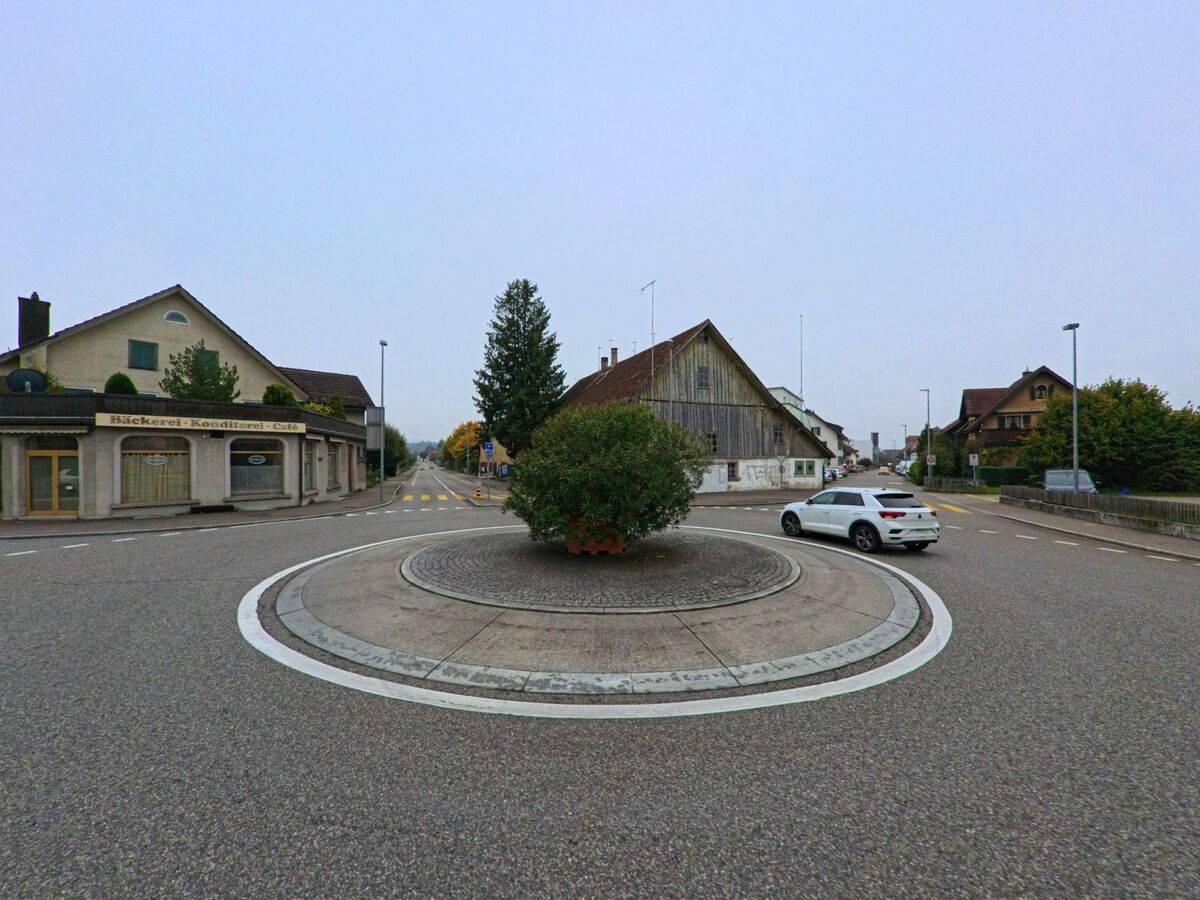 Ein kleiner Kreisverkehr mit bepflanzter Mittelinsel liegt im Zentrum von Fislisbach (5442, Kanton Aargau), umgeben von Wohnhäusern und einer Bäckerei mit Café.