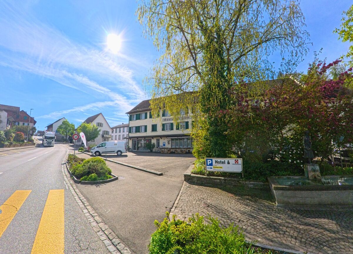 In der Gemeinde Flaach, PLZ 8416 im Kanton Zürich, fällt Sonnenlicht auf das Gebäude eines Hotels an der Hauptstrasse mit angrenzendem Parkplatz und einer kleinen Brunnenanlage im Vordergrund.