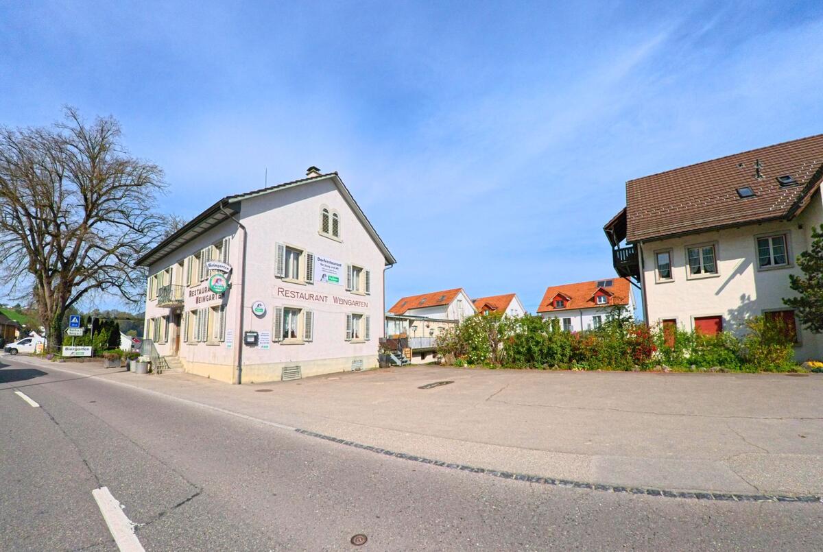 Im Zentrum von Flaach, PLZ 8416 im Kanton Zürich, steht das helle Gebäude des Restaurants Weingarten an einer Hauptstrasse, umgeben von Wohnhäusern mit roten Ziegeldächern.