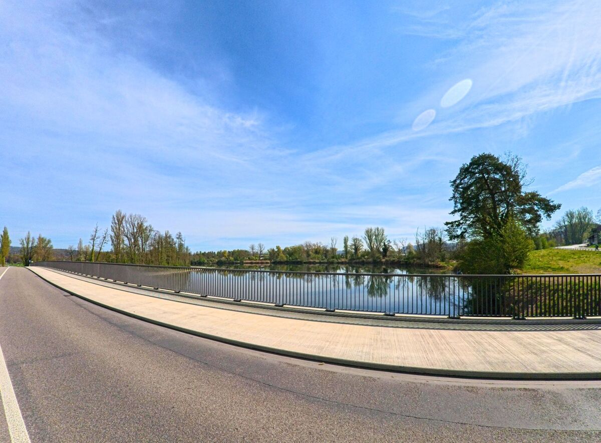 In Flaach, PLZ 8455 im Kanton Zürich, führt eine moderne Strassenbrücke mit Metallgeländer über einen ruhigen Fluss, gesäumt von Bäumen und Wiesen.